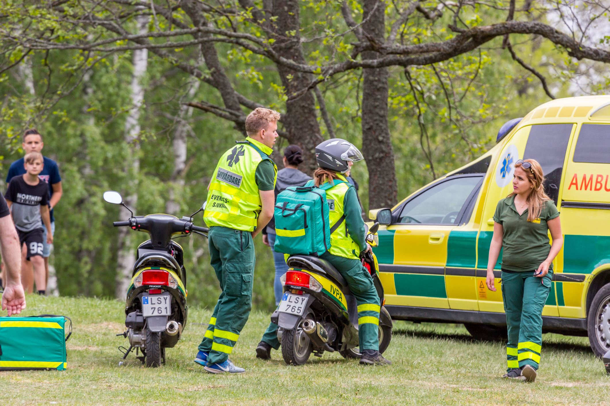 Stockholm, Sweden - June 03, 2017: Rear view of three pramedics and different ambulance vehicles outdoors, non-emergency rendezvous at public park. Female paramedic sitting on motorscooter in front of ambulance, civil man and boy standing beside.