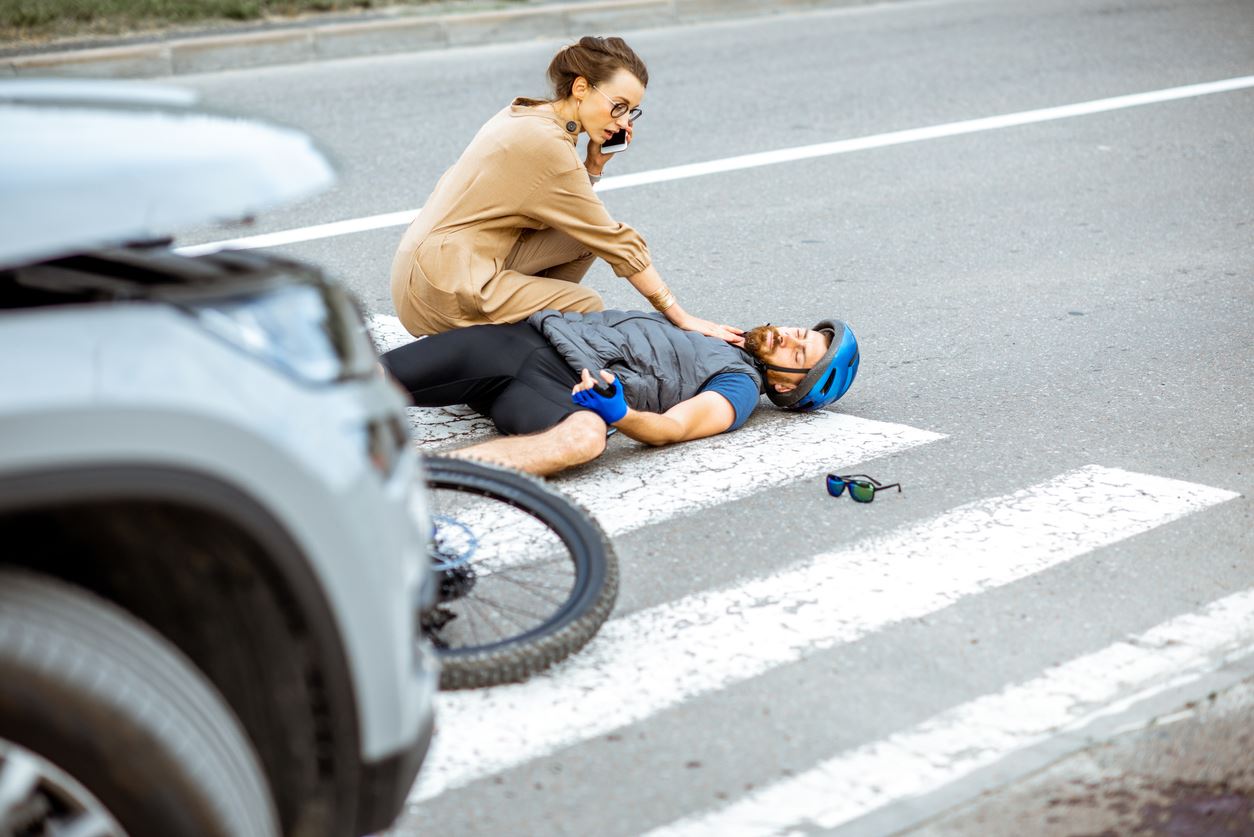 Road accident with injured cyclist lying on the pedestrian crossing near the broken bicycle, worried woman driver calling and checking men's pulse (Road accident with injured cyclist lying on the pedestrian crossing near the broken bicycle, worried wo S